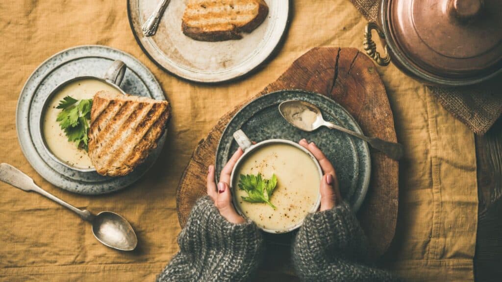 A person holds a mug of creamy soup garnished with herbs, next to a plate of grilled bread on a rustic table setting with spoons and a pot visible.