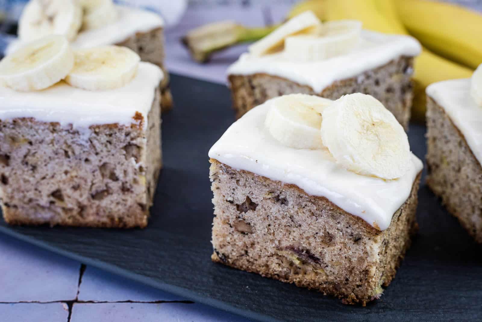 Squares of banana cake topped with white frosting and banana slices are arranged on a dark serving tray, with bananas visible in the background.