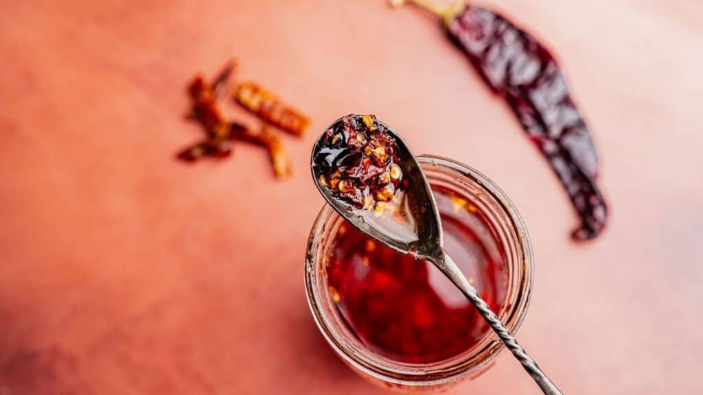 A spoon holding chili oil and chili crisp is held above an open jar, with dried chili peppers scattered on a pinkish surface nearby.