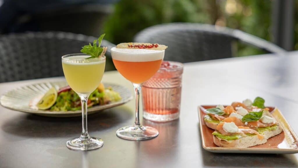 Two cocktails and a glass of water sit on a table next to plates of avocado toast with salmon and a salad, with patio chairs and greenery in the background.