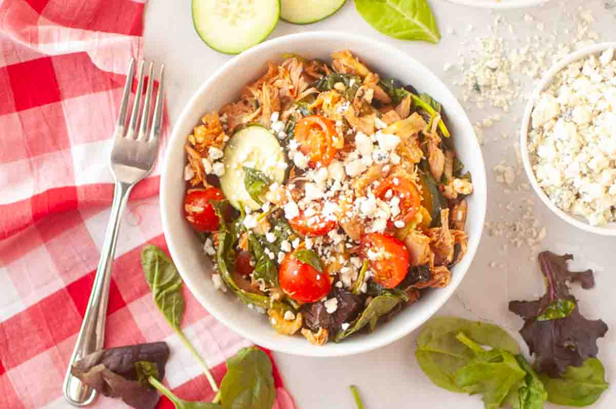 A bowl of salad with shredded chicken, grape tomatoes, cucumber, leafy greens, and crumbled cheese sits on a white surface next to a fork.