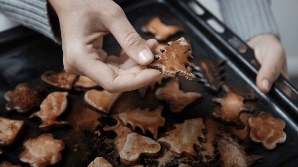 A person holds a tree-shaped cookie above a baking tray filled with assorted baked cookies in various shapes.