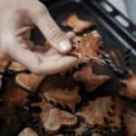 A person holds a tree-shaped cookie above a baking tray filled with assorted baked cookies in various shapes.