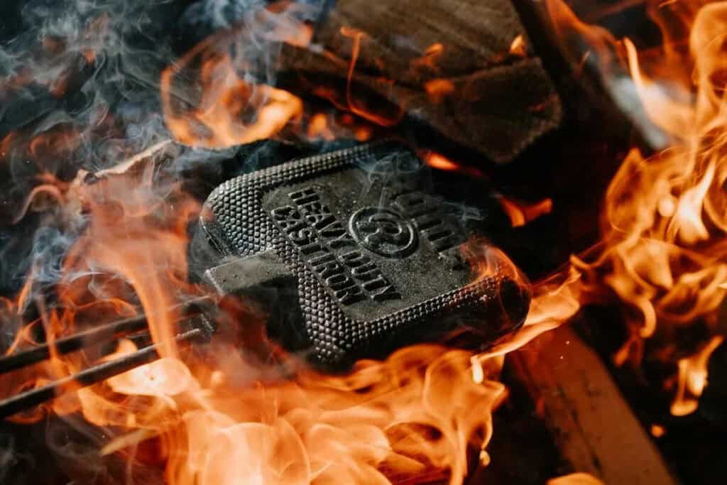 A heavy-duty cast iron sandwich press is heated in an open flame, surrounded by burning wood and fire&mdash;perfect for outdoor camping menus.