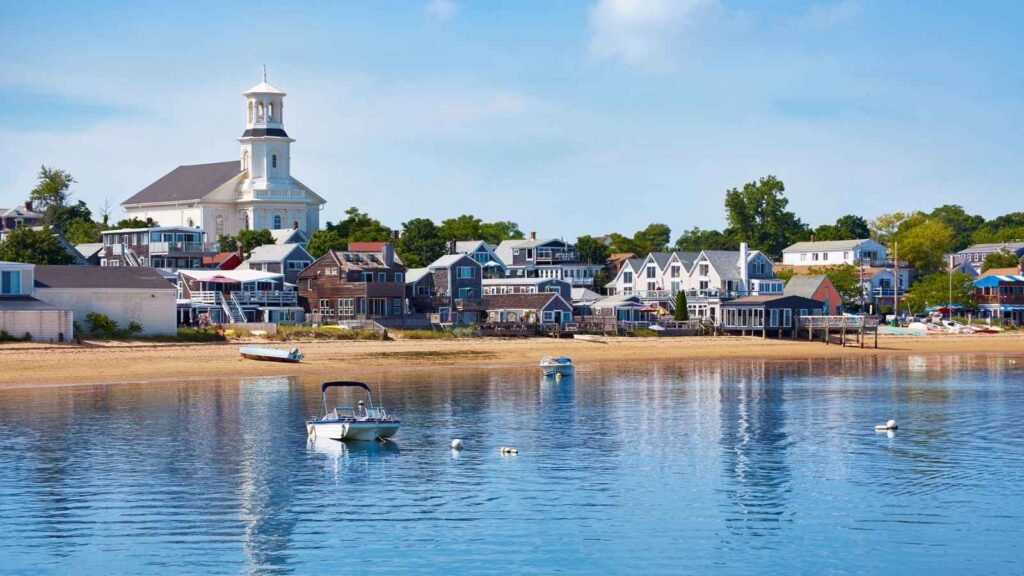 Small boats float near the sandy shore of a coastal town with colorful houses and a large white church under a blue sky.