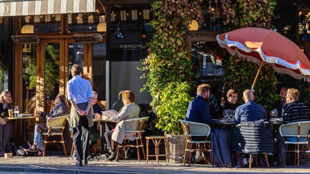 People are seated at outdoor tables of a restaurant, eating and talking. A waiter stands nearby, and there is greenery and an umbrella providing shade.