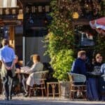 People are seated at outdoor tables of a restaurant, eating and talking. A waiter stands nearby, and there is greenery and an umbrella providing shade.
