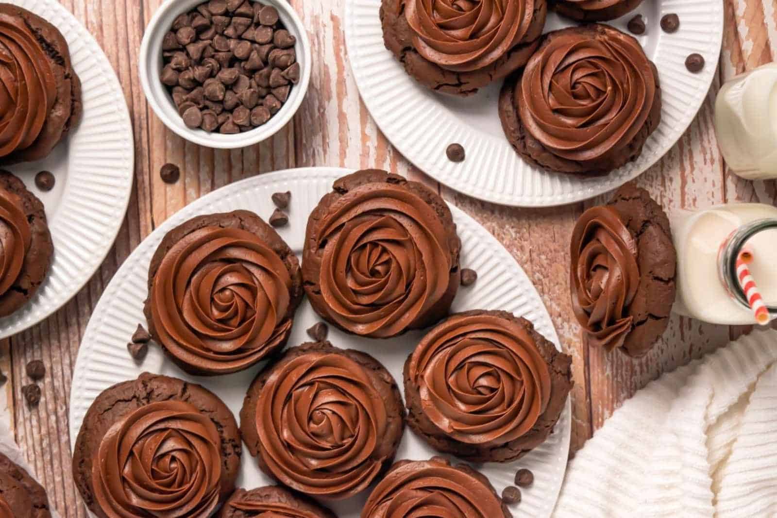 Plates of chocolate cookies with swirled chocolate frosting, a bowl of chocolate chips, and bottles of milk with striped straws on a wooden surface.