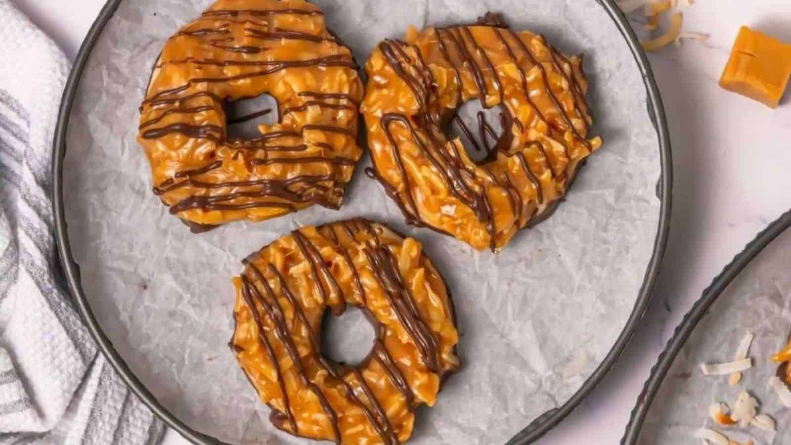 Three caramel and chocolate-drizzled cookies with coconut topping are arranged on a parchment-lined plate.