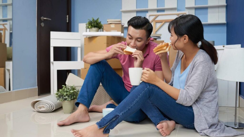 A couple sits on the floor among moving boxes, eating sandwiches and holding mugs, with unpacked household items around them.