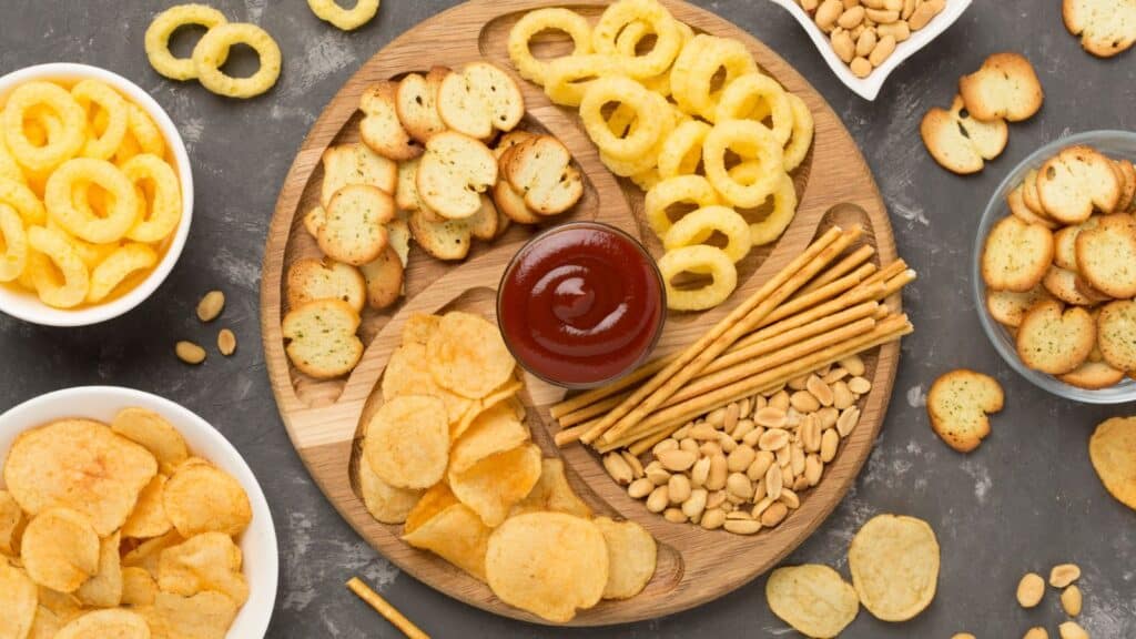 A wooden platter with potato chips, bread crisps, onion rings, grissini, peanuts, and a bowl of red dipping sauce, surrounded by bowls of chips, crisps, and peanuts.