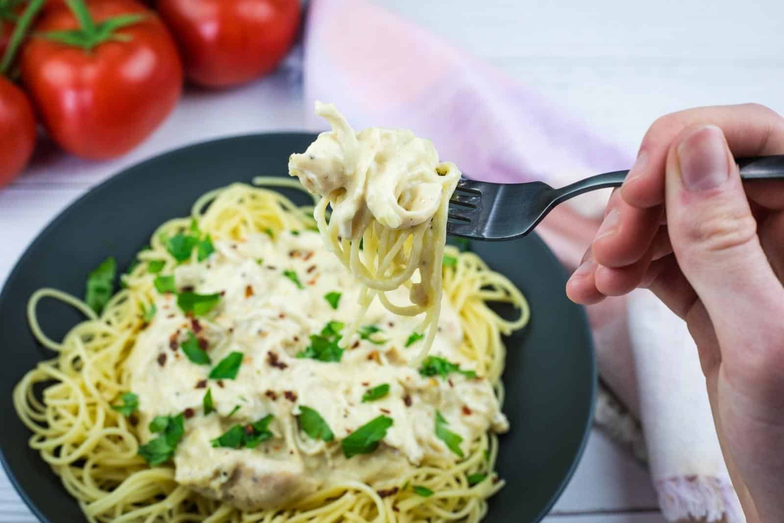 A hand holds a fork with a bite of creamy spaghetti above a black plate filled with pasta, topped with sauce and parsley. Fresh tomatoes sit in the background.