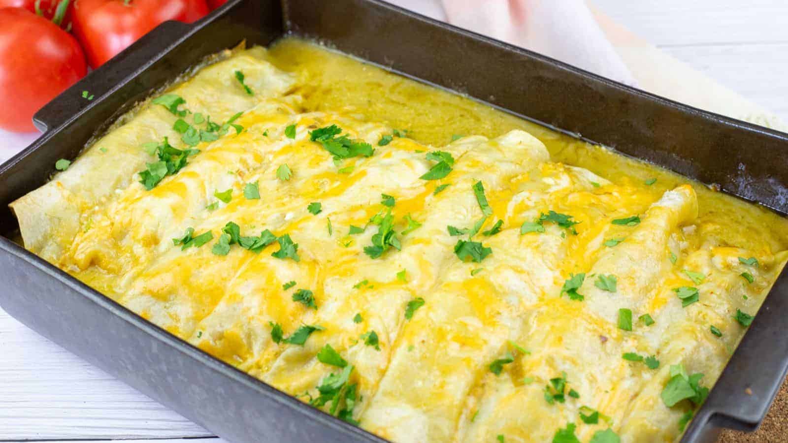 A baking dish filled with creamy green chili enchiladas topped with chopped cilantro, with tomatoes visible in the background.