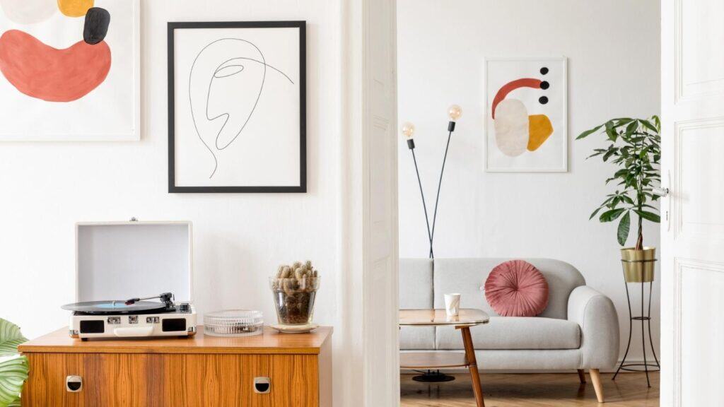 A modern living room with abstract wall art, a record player on a wooden cabinet, a small gray sofa with a pink pillow, a floor lamp, and a potted plant.