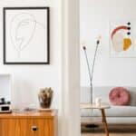 A modern living room with abstract wall art, a record player on a wooden cabinet, a small gray sofa with a pink pillow, a floor lamp, and a potted plant.