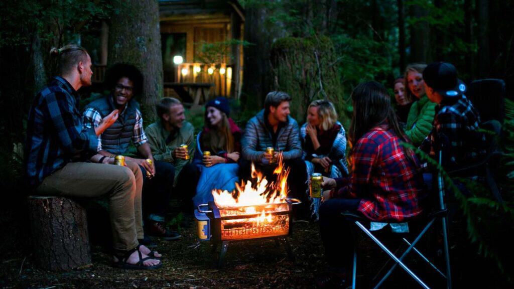 A group of people in casual clothes sit around a campfire in a wooded area, talking and holding drinks, enjoying one of their simpler trips, with a cabin visible in the background.