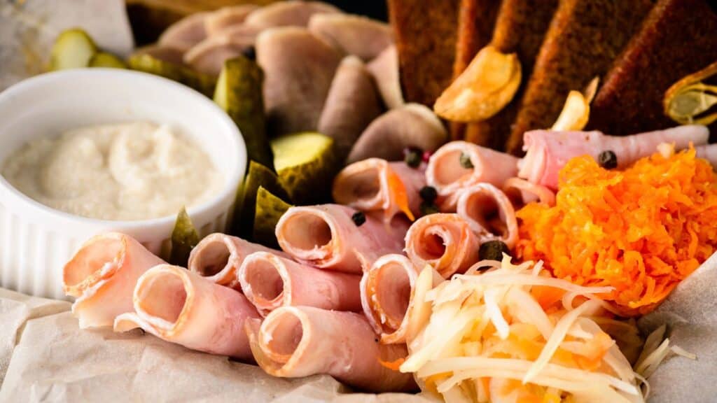 A beautiful snack board featuring rolled ham, sliced pickles, shredded carrots, sauerkraut, rye bread, garlic, and a small bowl of white dipping sauce.