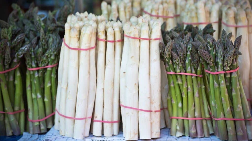 Bundles of white and green asparagus are displayed upright, held together with pink rubber bands, on a market stall.
