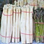 Bundles of white and green asparagus are displayed upright, held together with pink rubber bands, on a market stall.