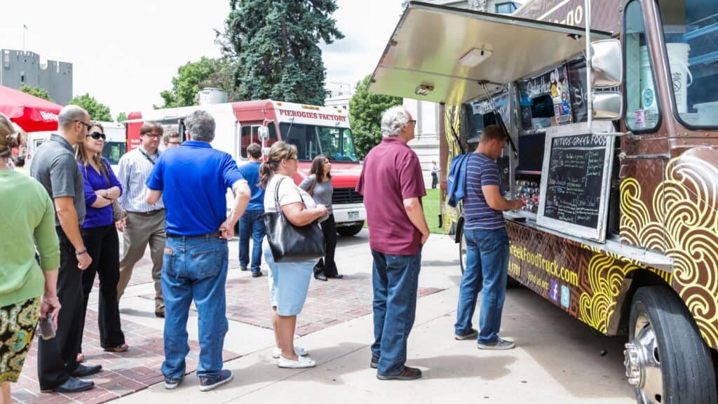 People stand in line at food trucks parked on a sidewalk, waiting to order food on a sunny day.