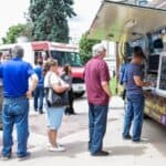 People stand in line at food trucks parked on a sidewalk, waiting to order food on a sunny day.