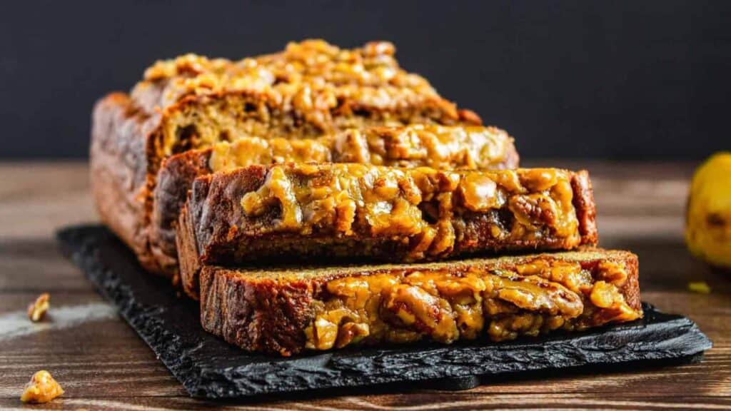 A loaf of banana bread topped with chopped walnuts, partially sliced, sits on a black slate serving board against a dark background.
