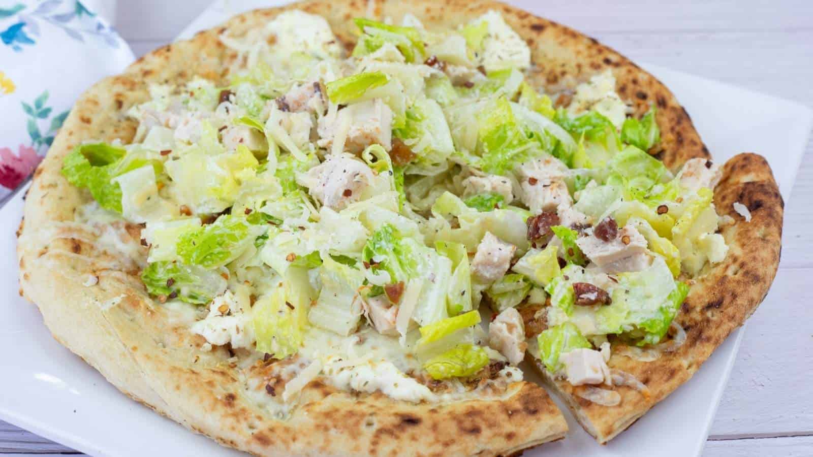 A pizza topped with chopped romaine lettuce, grilled chicken pieces, bacon bits, and Caesar dressing on a white plate.