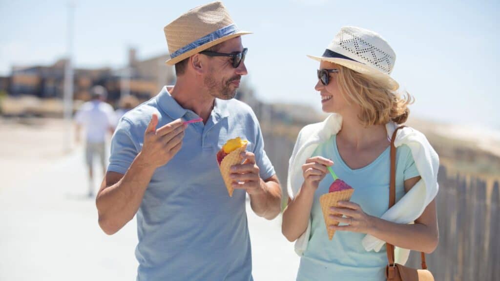 A man and a woman wearing hats and sunglasses eat ice cream cones while walking outdoors on a sunny day.