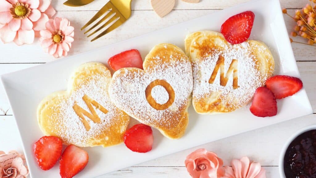 Three heart-shaped pancakes dusted with powdered sugar spell "MOM" on a white plate, garnished with sliced strawberries. Flowers and cutlery are arranged around the plate.