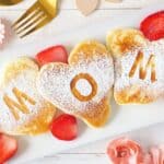 Three heart-shaped pancakes dusted with powdered sugar spell "MOM" on a white plate, garnished with sliced strawberries. Flowers and cutlery are arranged around the plate.