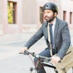 A man in a suit and helmet rides a bicycle outdoors, carrying a messenger bag.