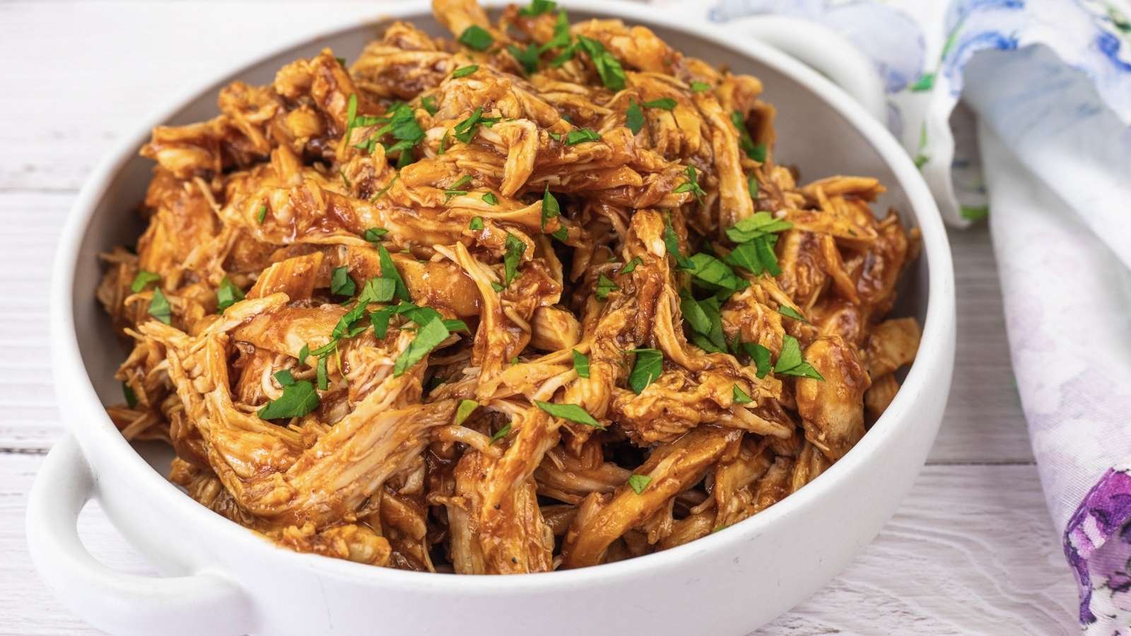 Slow Cooker BBQ Chicken garnished with chopped parsley in a white bowl on a light wooden surface.