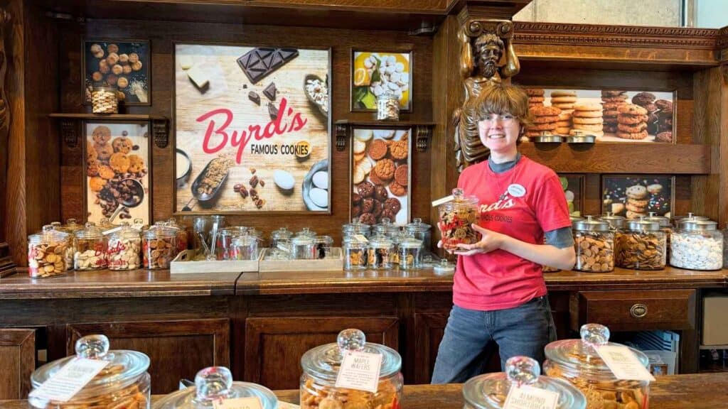 A person in a red shirt stands behind a wooden counter with jars of cookies at Byrd's Famous Cookies shop in Savannah, surrounded by cookie displays and signage.