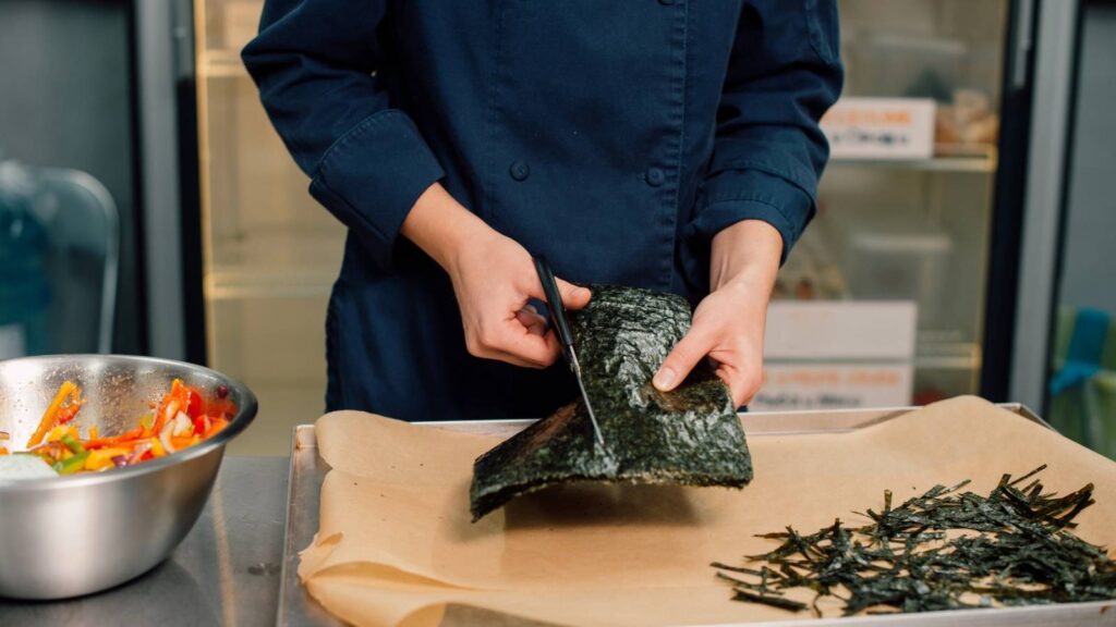 A person in a blue chef coat cuts sheets of dried seaweed on parchment paper, with a bowl of sliced vegetables nearby.