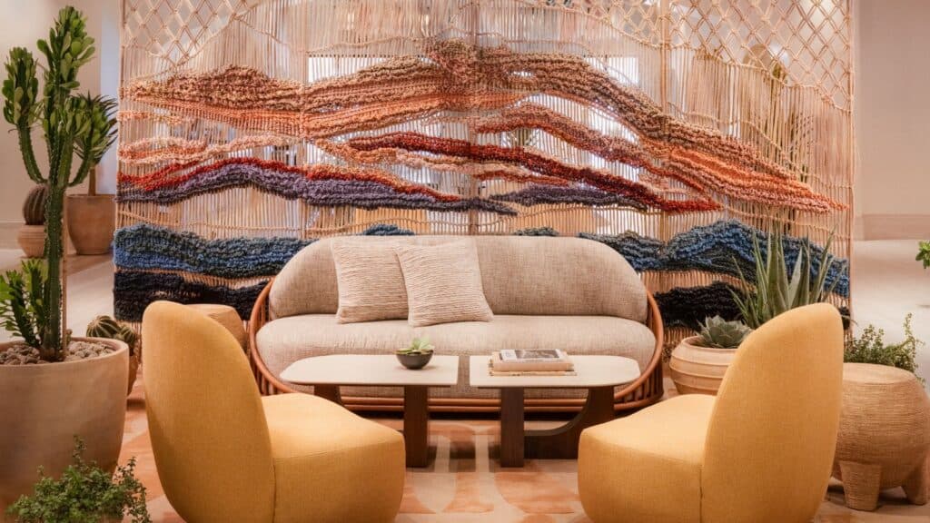 A modern lounge area with a beige sofa, two yellow chairs, a wooden coffee table, and a colorful woven wall hanging, surrounded by potted plants.