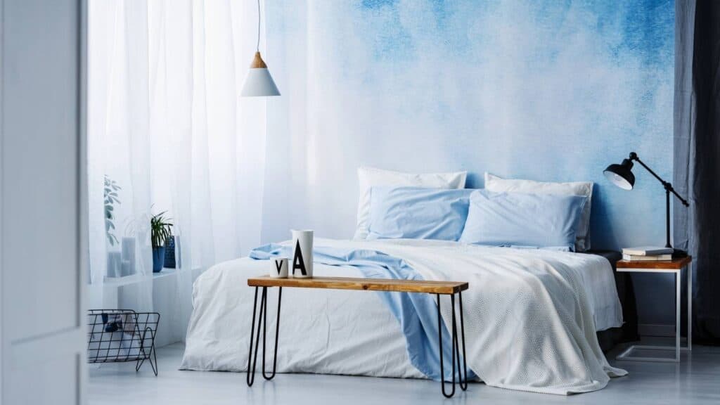 Modern bedroom with a blue and white color scheme, featuring a bed with light blue bedding, a wooden bench, and minimalist decor.