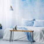 Modern bedroom with a blue and white color scheme, featuring a bed with light blue bedding, a wooden bench, and minimalist decor.