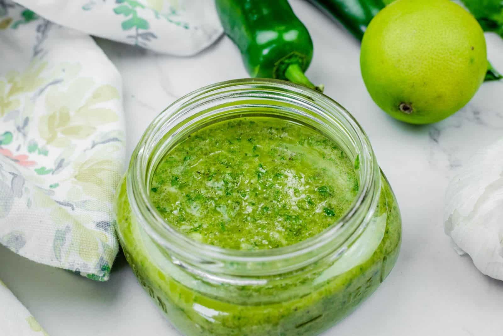 An open glass jar filled with Lime-Garlic Cilantro Marinade sits on a white surface next to a green chili, a lime, a garlic bulb, and a patterned cloth.