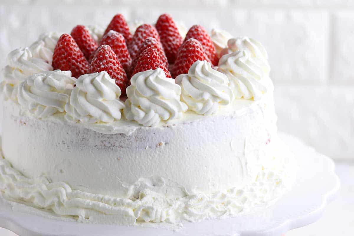 A round cake covered in white whipped cream, topped with whole strawberries and swirls of whipped cream, displayed on a white cake stand.