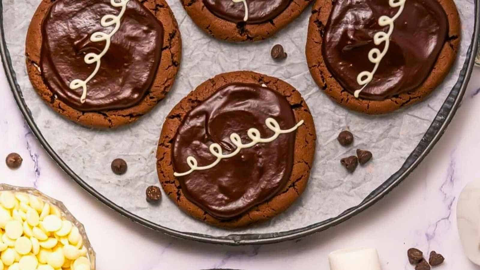 Four large chocolate cookies with glossy chocolate icing and white swirl decoration are arranged on a round tray lined with parchment paper. White chocolate chips are visible nearby.