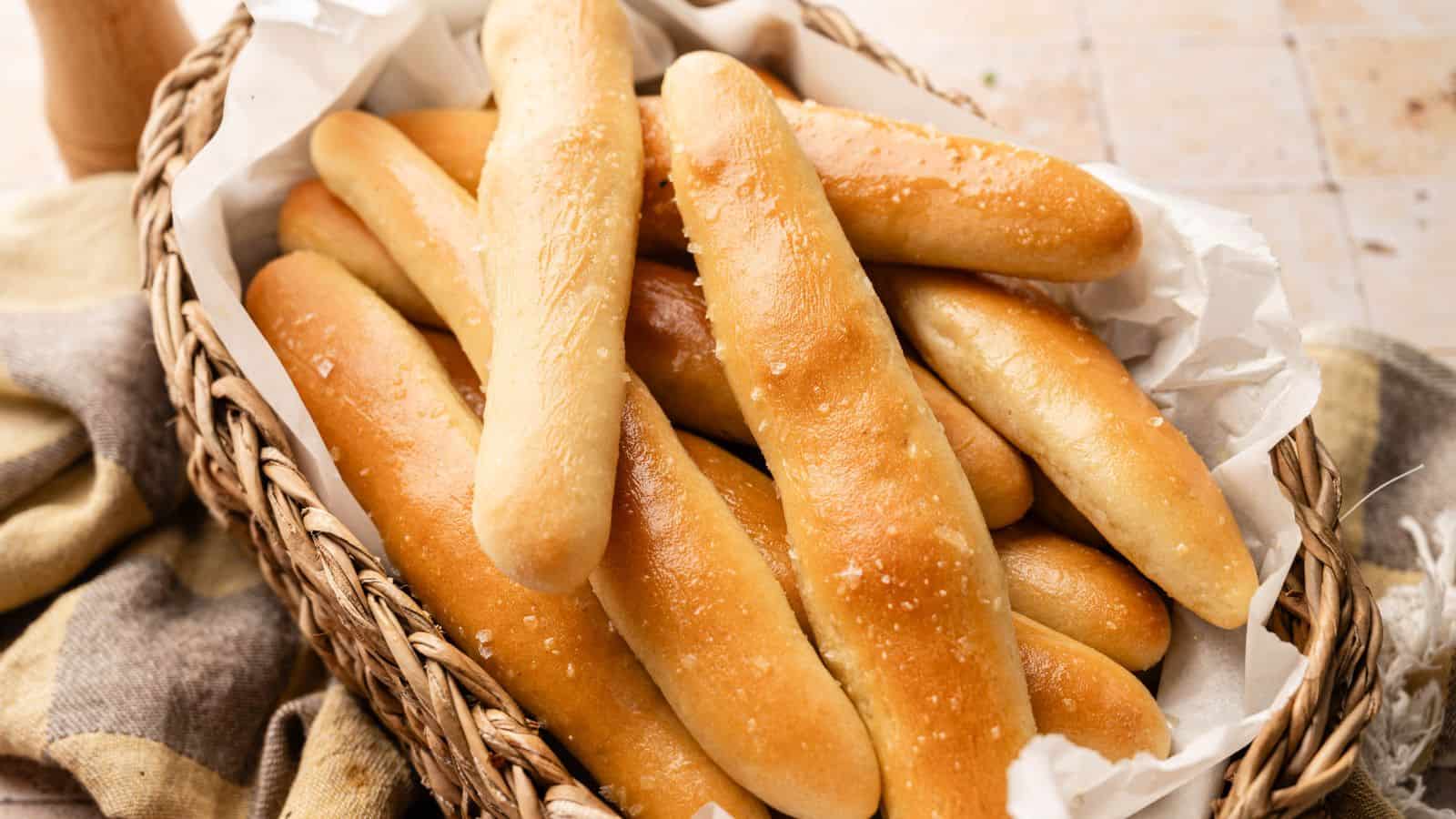A wicker basket lined with parchment paper holds several golden-brown breadsticks sprinkled with coarse salt.
