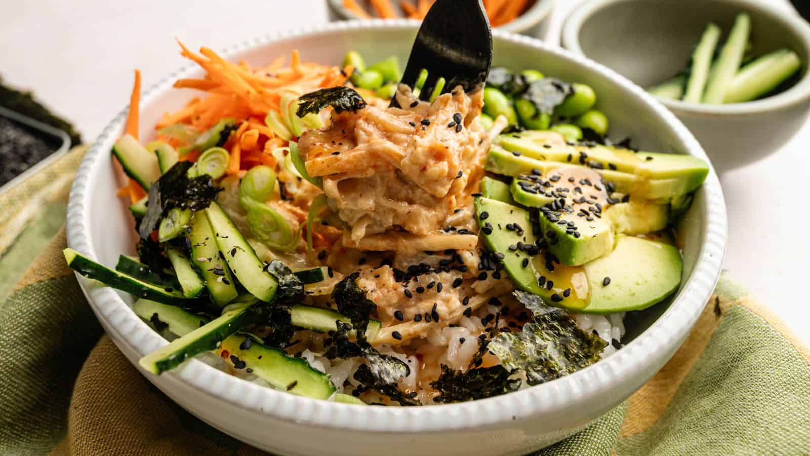 A bowl of rice topped with shredded carrots, cucumber, avocado, edamame, nori, black sesame seeds, and a creamy sauce, with a fork lifting a bite.