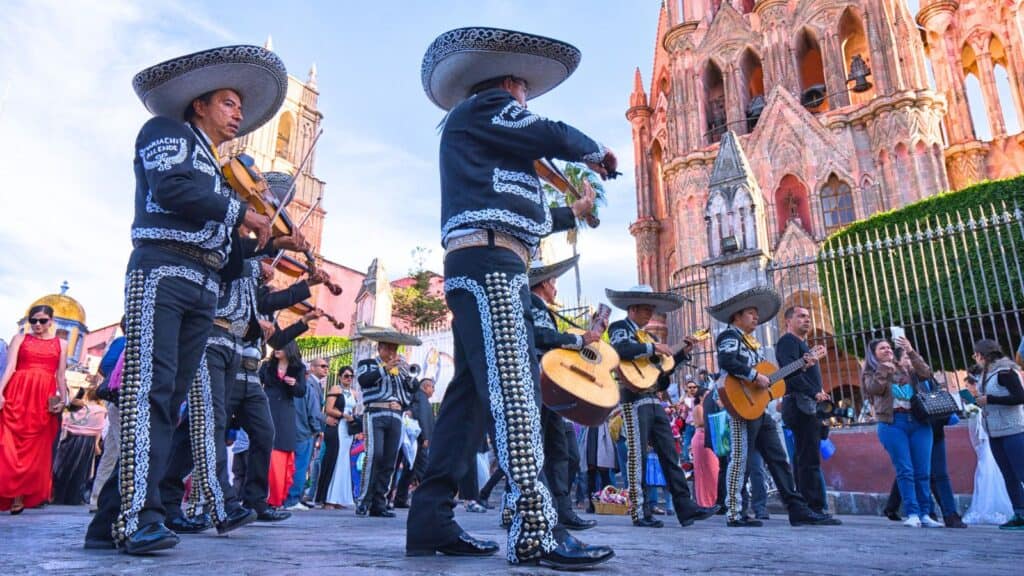 A mariachi band performs in a public square with an ornate church and onlookers in the background during daylight, capturing the vibrant spirit of a Central Mexico vacation.