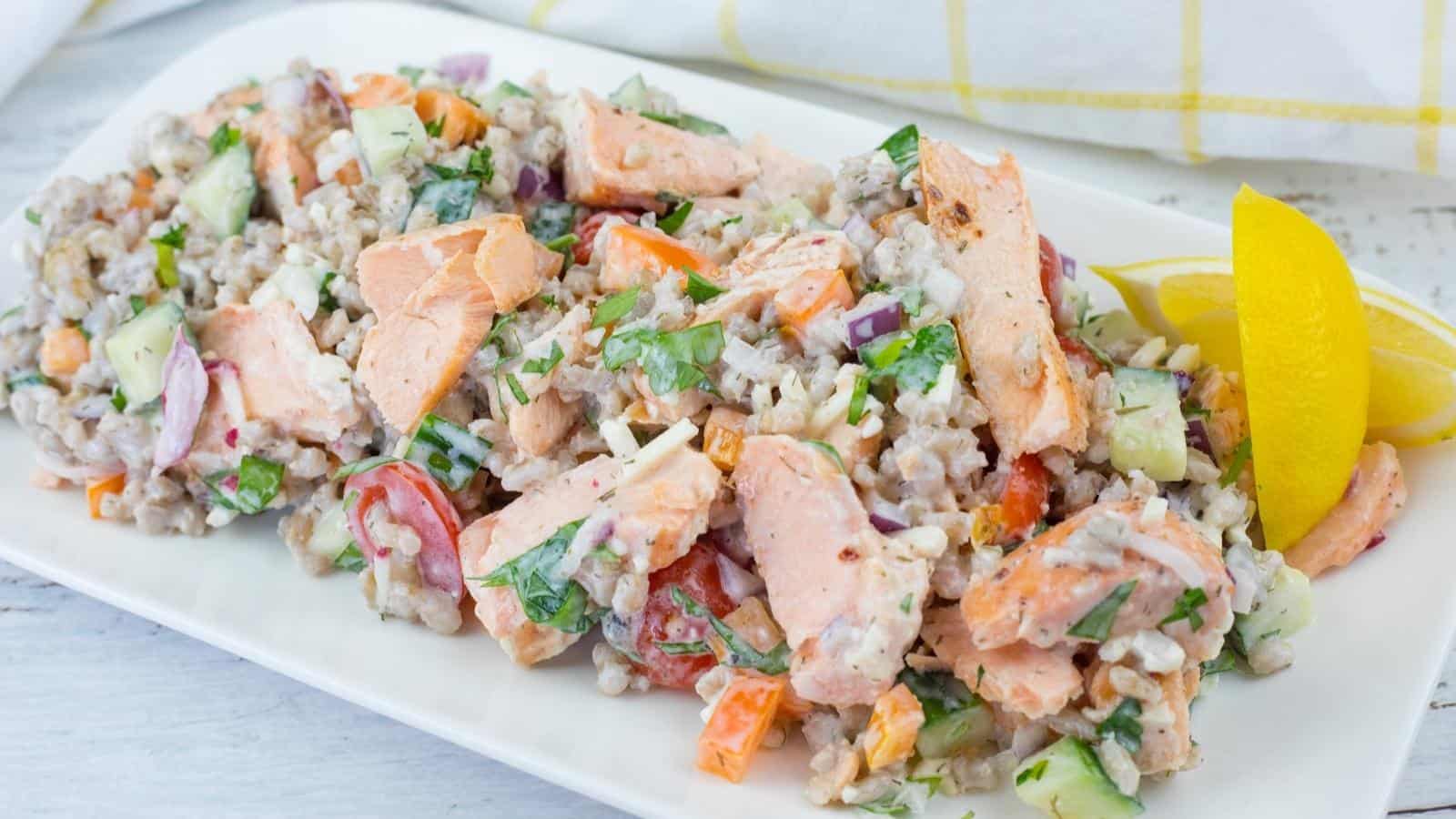 A rectangular white plate of Mediterranean Salmon Salad with Barley, topped with fresh herbs, and a lemon wedge on the side.