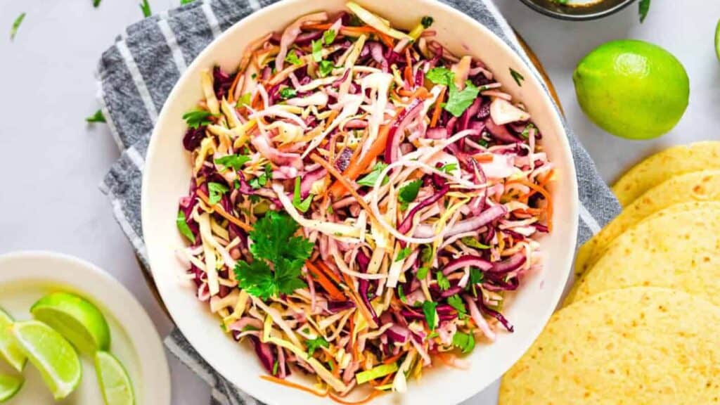 A bowl of colorful coleslaw with shredded cabbage, carrots, and cilantro sits on a striped towel next to lime wedges and tortillas.