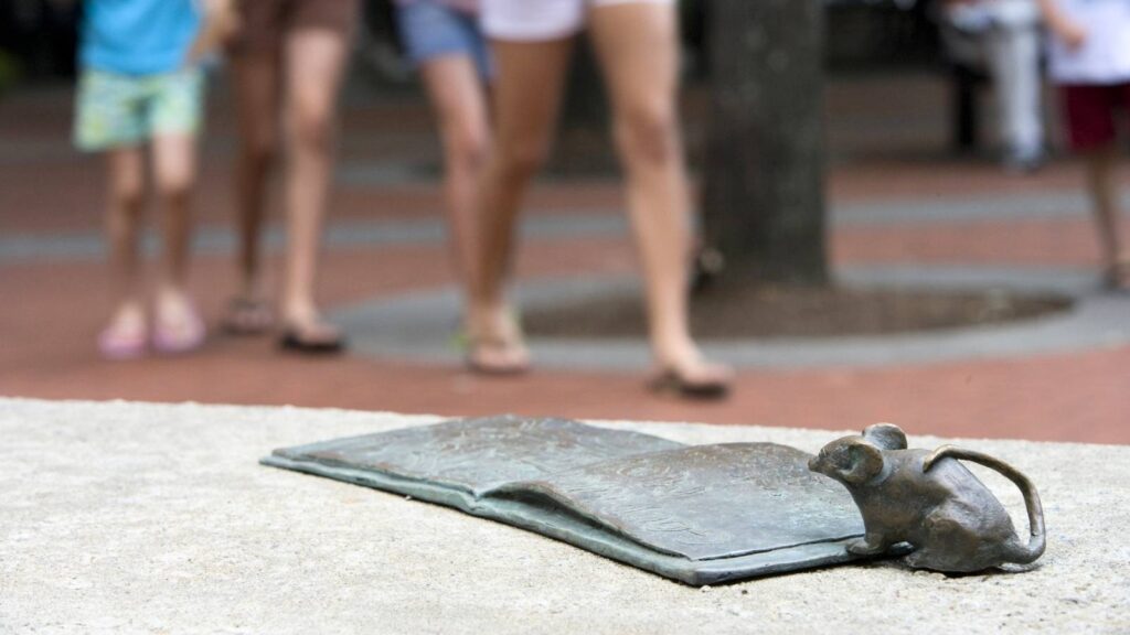 A small bronze sculpture of a mouse and an open book sits on a stone surface, with people walking in the blurred background&mdash;an inspiring scene to celebrate World Art Day.