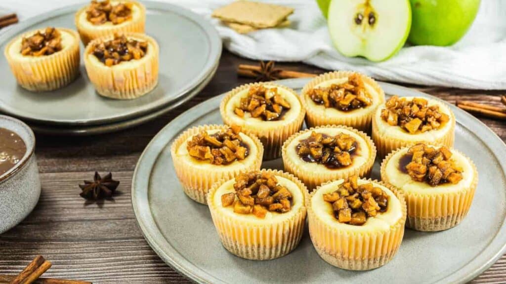Mini cheesecakes topped with diced caramelized apples are arranged on two plates, with star anise, a green apple, and graham crackers in the background.