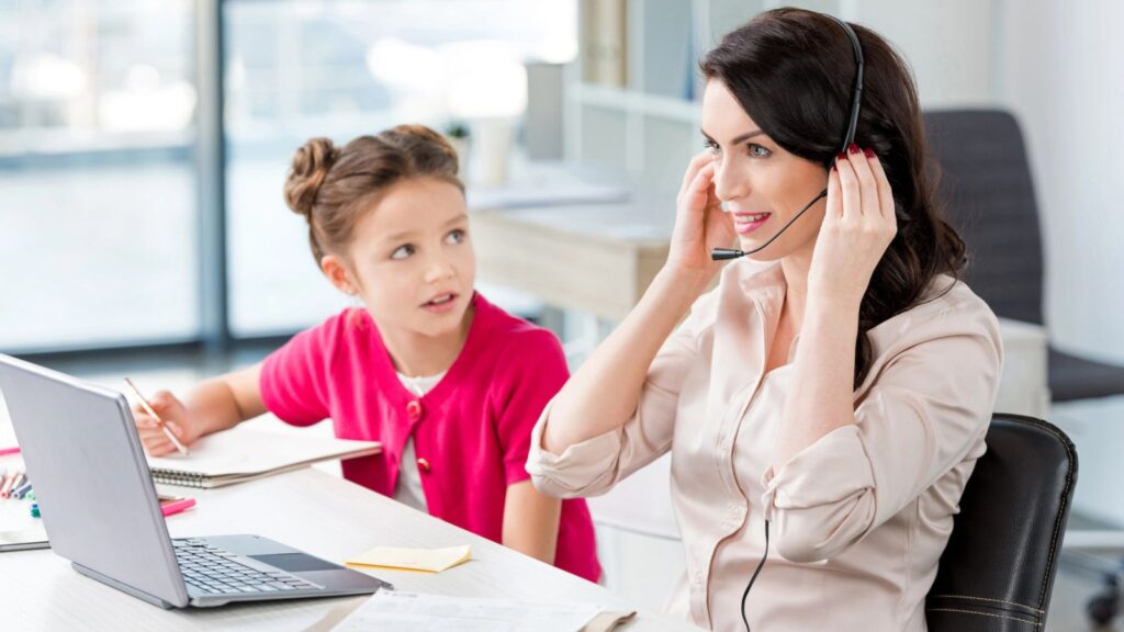 Woman wearing a headset works on a laptop at a desk while a young girl in a pink cardigan sits beside her, looking at her and holding a notebook.