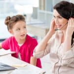 Woman wearing a headset works on a laptop at a desk while a young girl in a pink cardigan sits beside her, looking at her and holding a notebook.