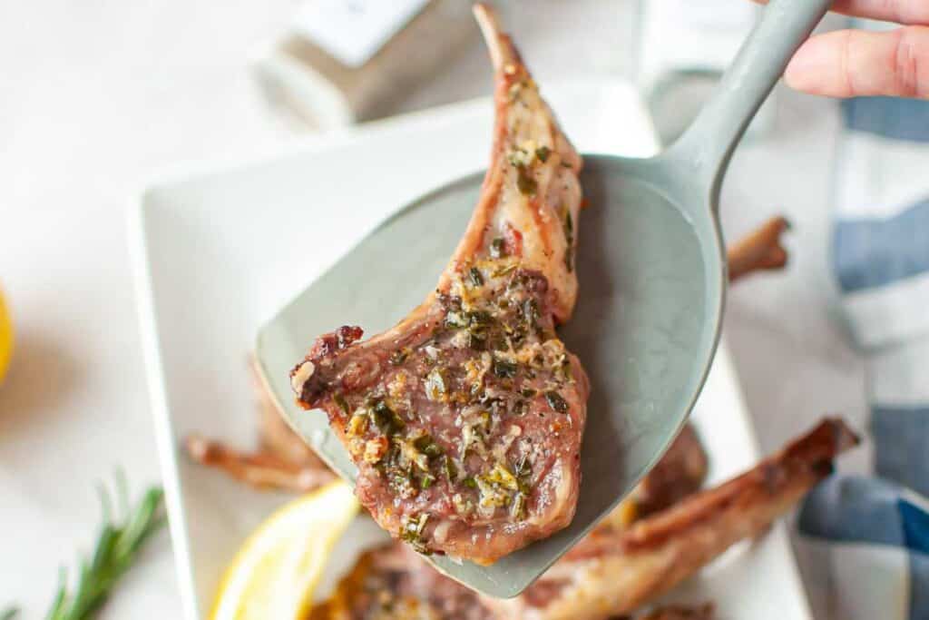 A close-up of a cooked lamb chop with herbs, held on a spatula above a white plate with more lamb chops and lemon slices.